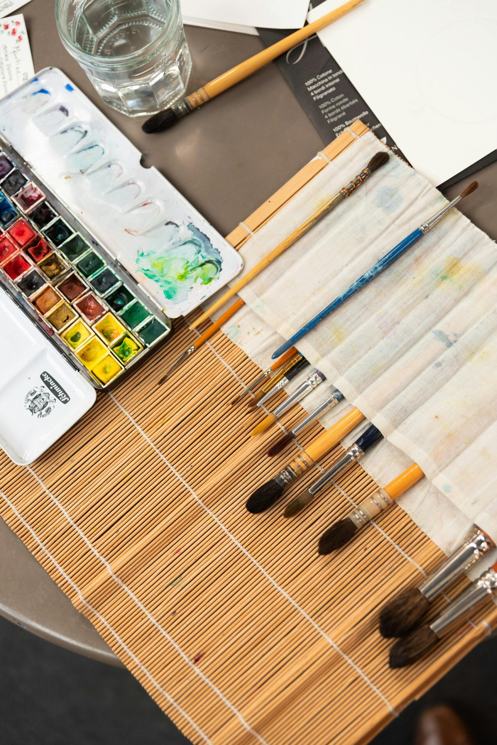A detailed view of an artist's workspace featuring watercolor paints and brushes on a bamboo mat.